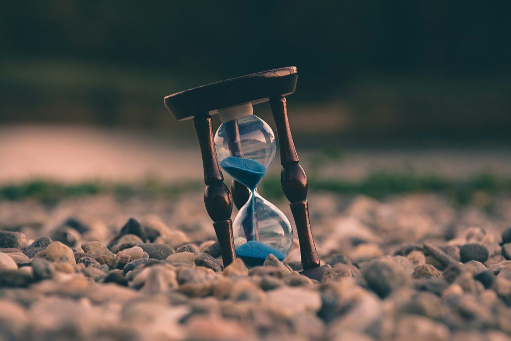 Image of an hourglass with blue sand, half in the top an half below, sitting askew in a rocky ground.
