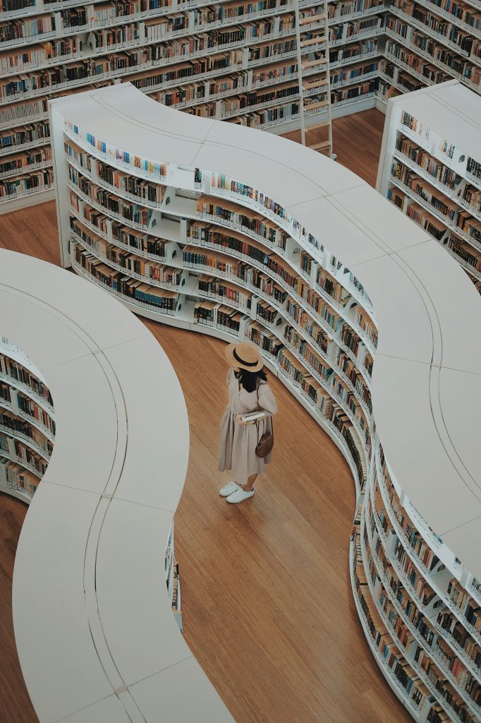 Photo of library stacks with curved shelving and a woman dressed in white with a hat browsing. 