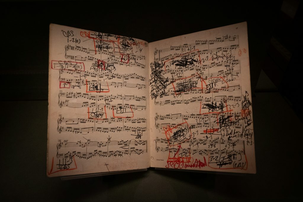 Photo of a book of music with two pages of musical notes shown in black with  black and red edits scribbled in handwriting on top.